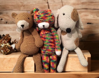 May include: Three crocheted stuffed toys: a brown bear, a rainbow-coloured cat, and a beige dog. The toys are sitting on a wooden shelf.