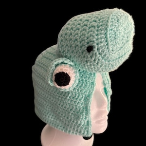 Może przedstawiać: Jasnoniebieska czapka z szydełka w kształcie żaby, z dużymi, okrągłymi oczami i wystającą głową. Czapka jest prezentowana na białej głowie manekina na czarnym tle. Oczy żaby są białe z czarnymi źrenicami.