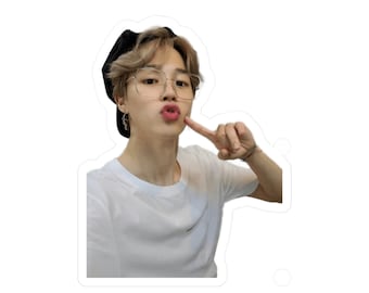 May include: A young person with brown hair wearing a black cap, clear framed glasses, and a white t-shirt. They are making a kissy face and pointing to their cheek.