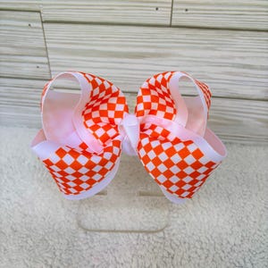 May include: A hair bow featuring a vibrant orange and white checkered pattern on a grosgrain ribbon. The bow is constructed with multiple loops of ribbon, creating a voluminous, layered appearance with a central knot securing the structure.