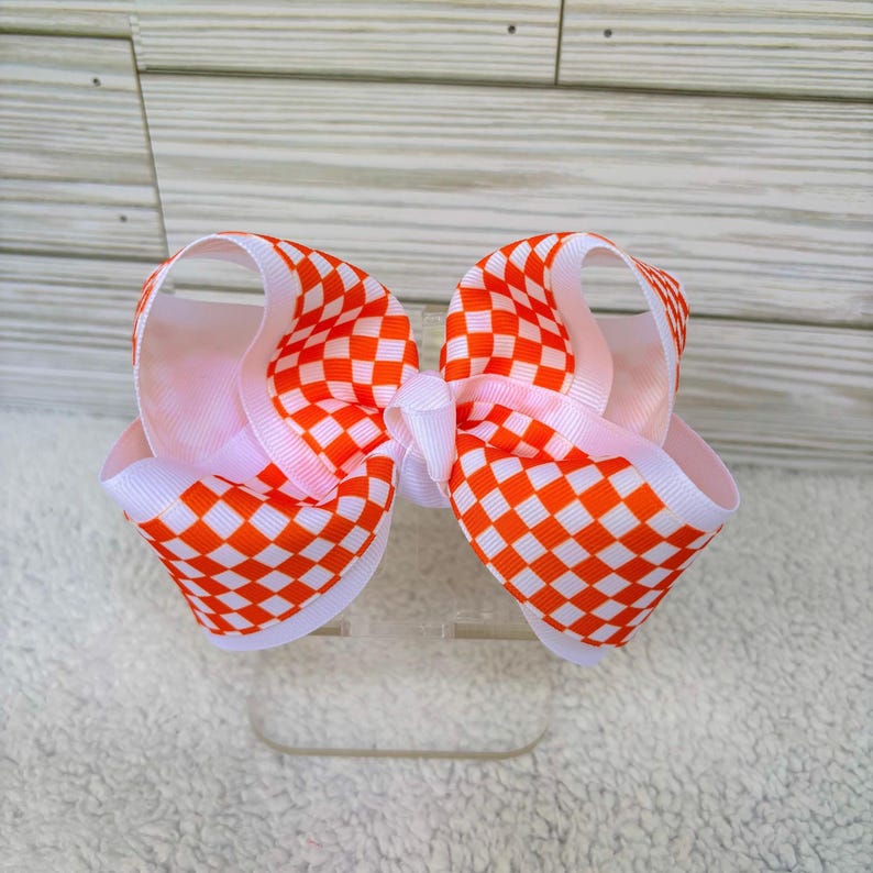 May include: A hair bow featuring a vibrant orange and white checkered pattern on a grosgrain ribbon. The bow is constructed with multiple loops of ribbon, creating a voluminous, layered appearance with a central knot securing the structure.