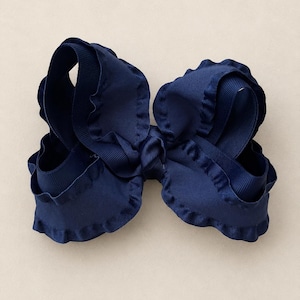 May include: A navy blue hair bow constructed from layered grosgrain ribbon. The bow features a central knot and wide, ruffled edges along the loops, creating a voluminous, multi-dimensional appearance with a consistent deep blue color throughout.