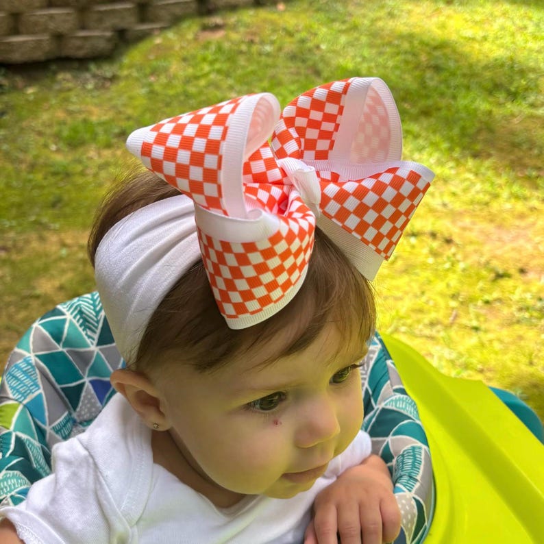 May include: A large hair bow featuring an orange and white checkered pattern attached to a plain white fabric headband. The bow is constructed from wide grosgrain ribbon with crisp, folded loops that create a voluminous, multi-layered appearance.