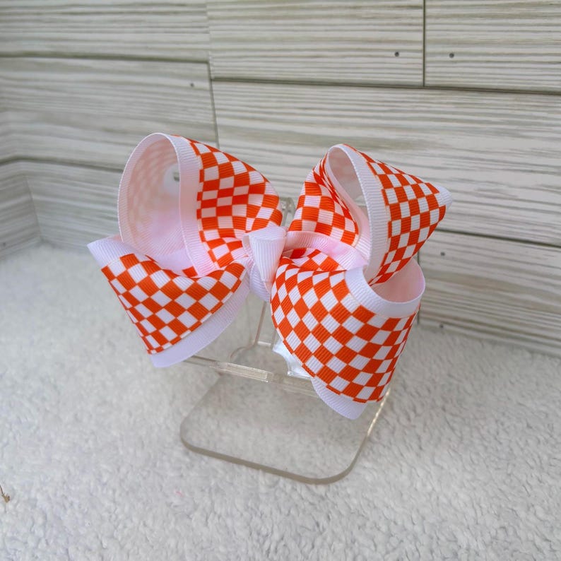 May include: A hair bow featuring a vibrant orange and white checkered pattern. The bow is constructed from wide grosgrain ribbon with crisp, folded loops and a central knot, showcasing a repeating square grid design throughout the fabric.