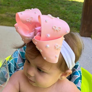 May include: A large light pink hair bow attached to a white elastic headband. The bow is made of ribbed fabric and is decorated with small white faux pearls and tiny white bow-shaped embellishments scattered across the surface of the fabric.