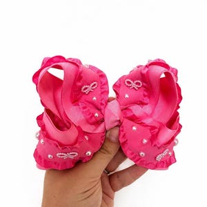 May include: A bright pink hair bow made of grosgrain ribbon with ruffled edges. The bow is decorated with small white faux pearls and tiny white bow-shaped embellishments scattered across the fabric surface, creating a textured appearance.