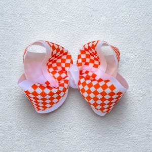 May include: A hair bow constructed from grosgrain ribbon featuring a vibrant orange and white checkered pattern. The bow is designed with multiple loops and a central knot, showcasing a structured, layered appearance with clean, crisp edges.
