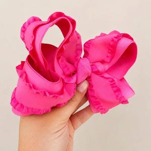 May include: A vibrant hot pink hair bow constructed from grosgrain ribbon. The accessory features multiple loops and layers with delicate ruffled edges along the perimeter of the ribbon, creating a voluminous and textured appearance.