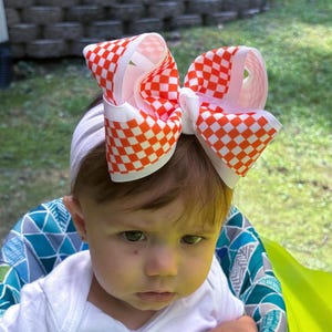 May include: A large hair bow featuring a vibrant orange and white checkered pattern. The bow is constructed from wide grosgrain ribbon with a central knot and is attached to a plain white elastic headband, creating a bold and structured accessory.