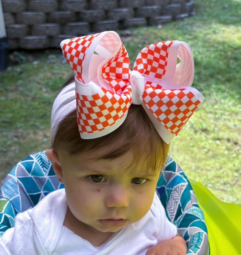 May include: A large hair bow featuring a vibrant orange and white checkered pattern. The bow is constructed from wide grosgrain ribbon with a central knot and is attached to a plain white elastic headband, creating a bold and structured accessory.