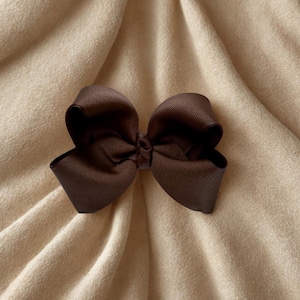 May include: A dark brown hair bow made of grosgrain ribbon. The bow is centered and has a classic bow shape. The background is a light beige fabric, creating a contrast with the bow's color.