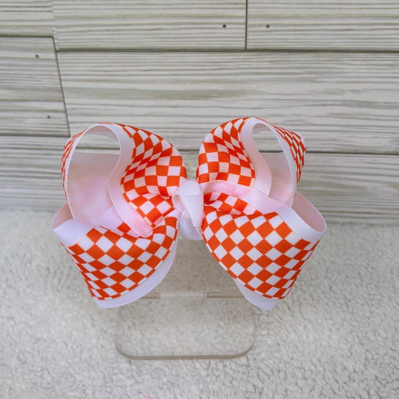 May include: A hair bow featuring a vibrant orange and white checkered pattern. The bow is constructed from layered grosgrain ribbon with a central knot detail, creating a voluminous, multi-looped structure with crisp, clean edges throughout.