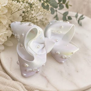 May include: A large white hair bow made of ribbed fabric, featuring a layered design adorned with scattered round white pearls and small clusters of tiny white beads attached to the surface of the loops, creating a textured and dimensional appearance.