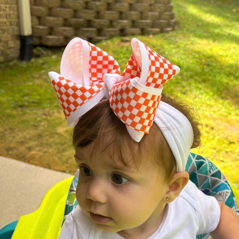 May include: A large hair bow featuring a vibrant orange and white checkered pattern, attached to a plain white elastic headband. The bow is constructed from wide grosgrain ribbon with crisp, folded loops that create a voluminous, multi-layered look.