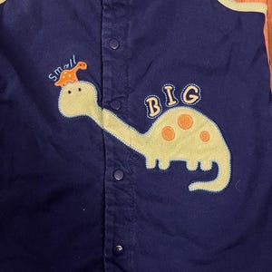 Puede incluir: Romper de beb&eacute; azul marino con una aplicaci&oacute;n de dinosaurio verde claro. El dinosaurio tiene manchas naranjas y est&aacute; etiquetado como "BIG". Encima del dinosaurio est&aacute; la palabra "small" con un dinosaurio a juego. El romper tiene una parte delantera abotonada y ribetes naranjas y verdes.