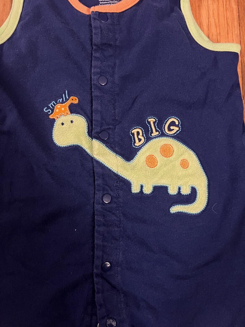 Puede incluir: Romper de beb&eacute; azul marino con una aplicaci&oacute;n de dinosaurio verde claro. El dinosaurio tiene manchas naranjas y est&aacute; etiquetado como "BIG". Encima del dinosaurio est&aacute; la palabra "small" con un dinosaurio a juego. El romper tiene una parte delantera abotonada y ribetes naranjas y verdes.