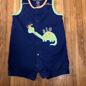 Puede incluir: Romper de beb&eacute; azul marino con ribete verde y naranja. El romper presenta una aplicaci&oacute;n de dinosaurio verde con las palabras "small" y "BIG". El romper tiene cierres de broche en la parte delantera y una etiqueta que dice "child of mine".