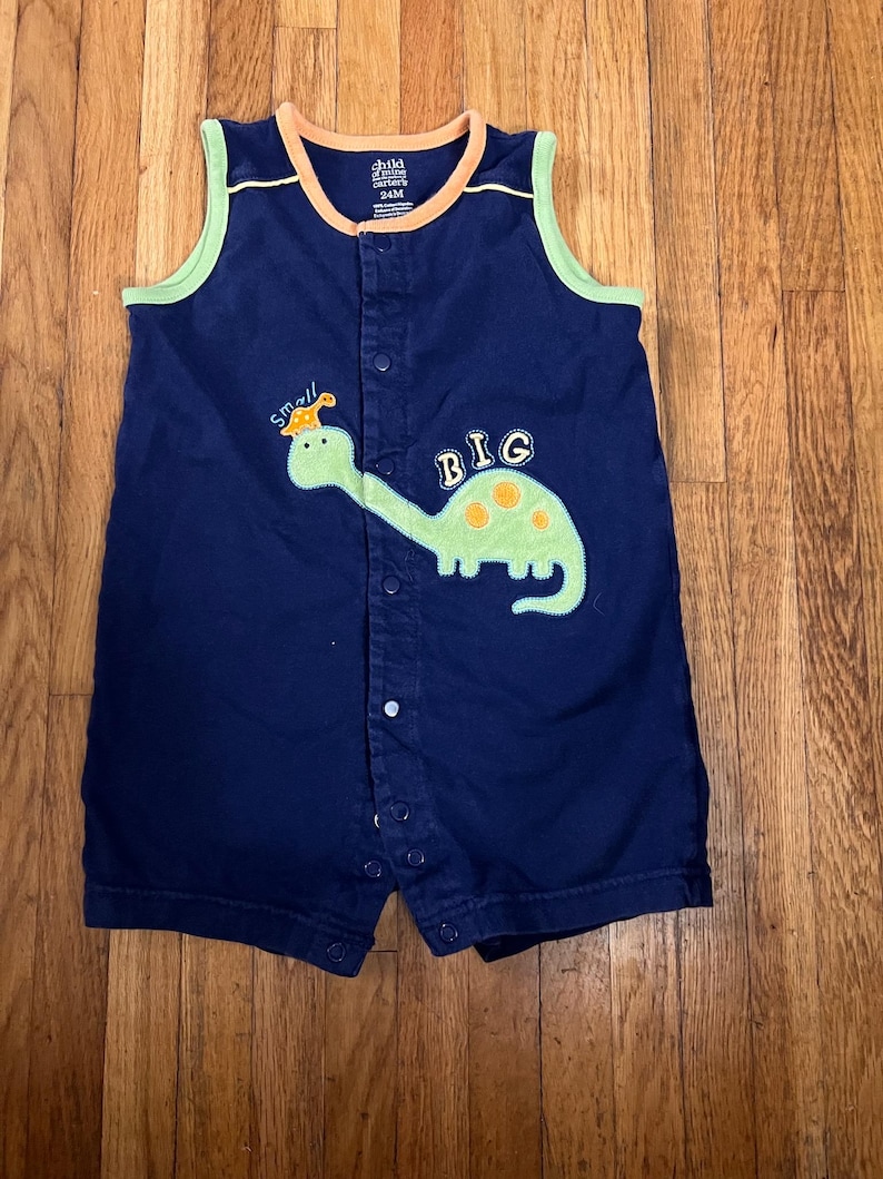 Puede incluir: Romper de beb&eacute; azul marino con ribete verde y naranja. El romper presenta una aplicaci&oacute;n de dinosaurio verde con las palabras "small" y "BIG". El romper tiene cierres de broche en la parte delantera y una etiqueta que dice "child of mine".