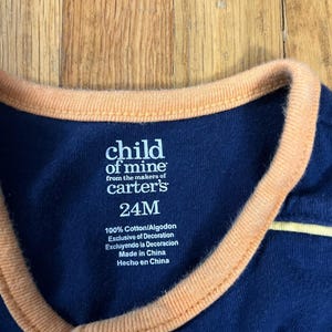 Puede incluir: Primer plano de un body de beb&eacute; azul marino con ribete naranja en el cuello. La etiqueta dice "child of mine from the makers of carter's 24M". El body est&aacute; hecho de 100% algod&oacute;n y es exclusivo de decoraci&oacute;n.