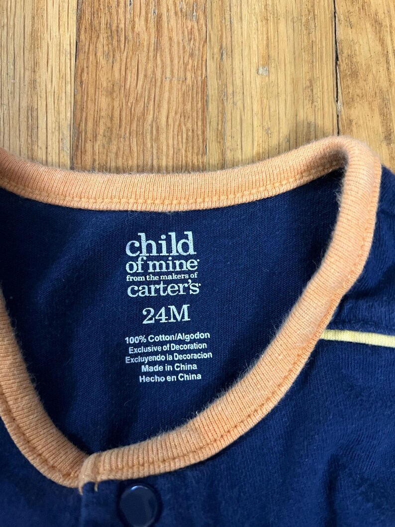 Puede incluir: Primer plano de un body de beb&eacute; azul marino con ribete naranja en el cuello. La etiqueta dice "child of mine from the makers of carter's 24M". El body est&aacute; hecho de 100% algod&oacute;n y es exclusivo de decoraci&oacute;n.