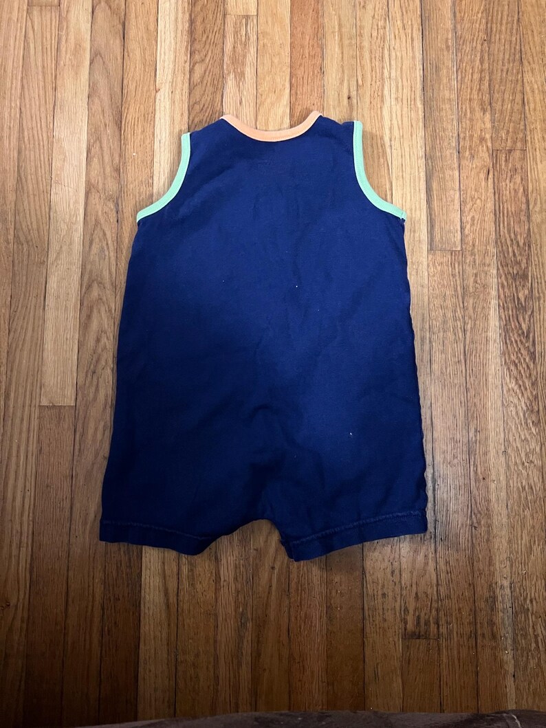 Puede incluir: Un pelele de beb&eacute; azul marino con cuello redondo y dise&ntilde;o sin mangas. El cuello y las sisas est&aacute;n ribeteados con un acento verde claro y naranja. El pelele est&aacute; hecho de una tela suave.