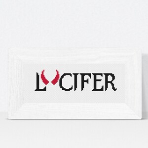 May include: A white framed cross-stitch with the word "LUCIFER" in black letters and red devil horns.