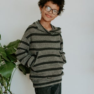 May include: A young person wearing a gray and black striped hooded sweatshirt with a front pocket. They are wearing black jeans and glasses.