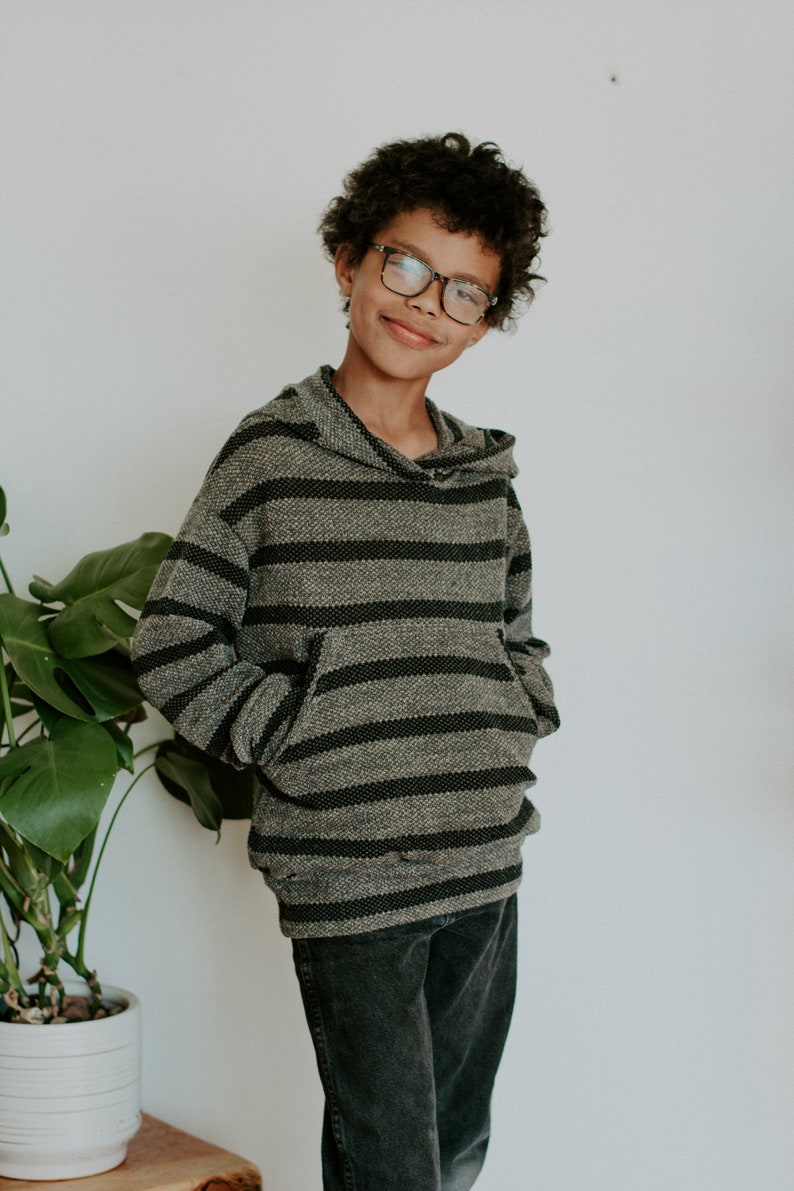 May include: A young person wearing a gray and black striped hooded sweatshirt with a front pocket. They are wearing black jeans and glasses.