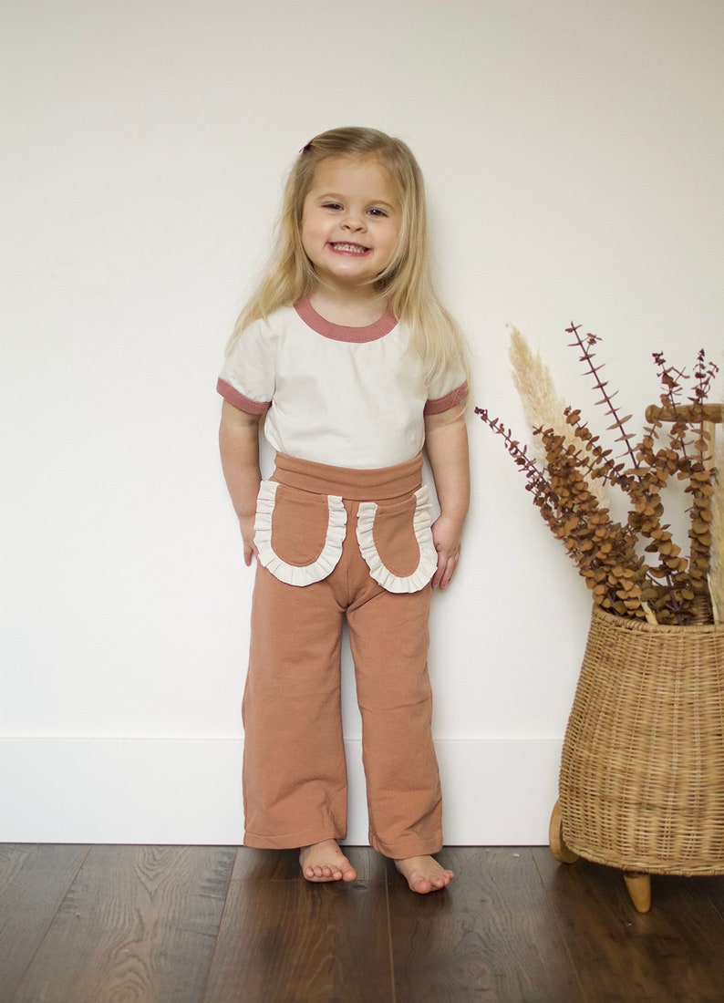 May include: A young girl wearing a white t-shirt with a pink trim and brown pants with white ruffle details. The pants have a wide leg style. The girl is smiling and standing in front of a white wall.