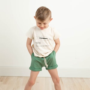 May include: A young boy wearing a cream colored t-shirt with a black and white illustration of a fish and the text "steelhead (steel-hed)" and green shorts.