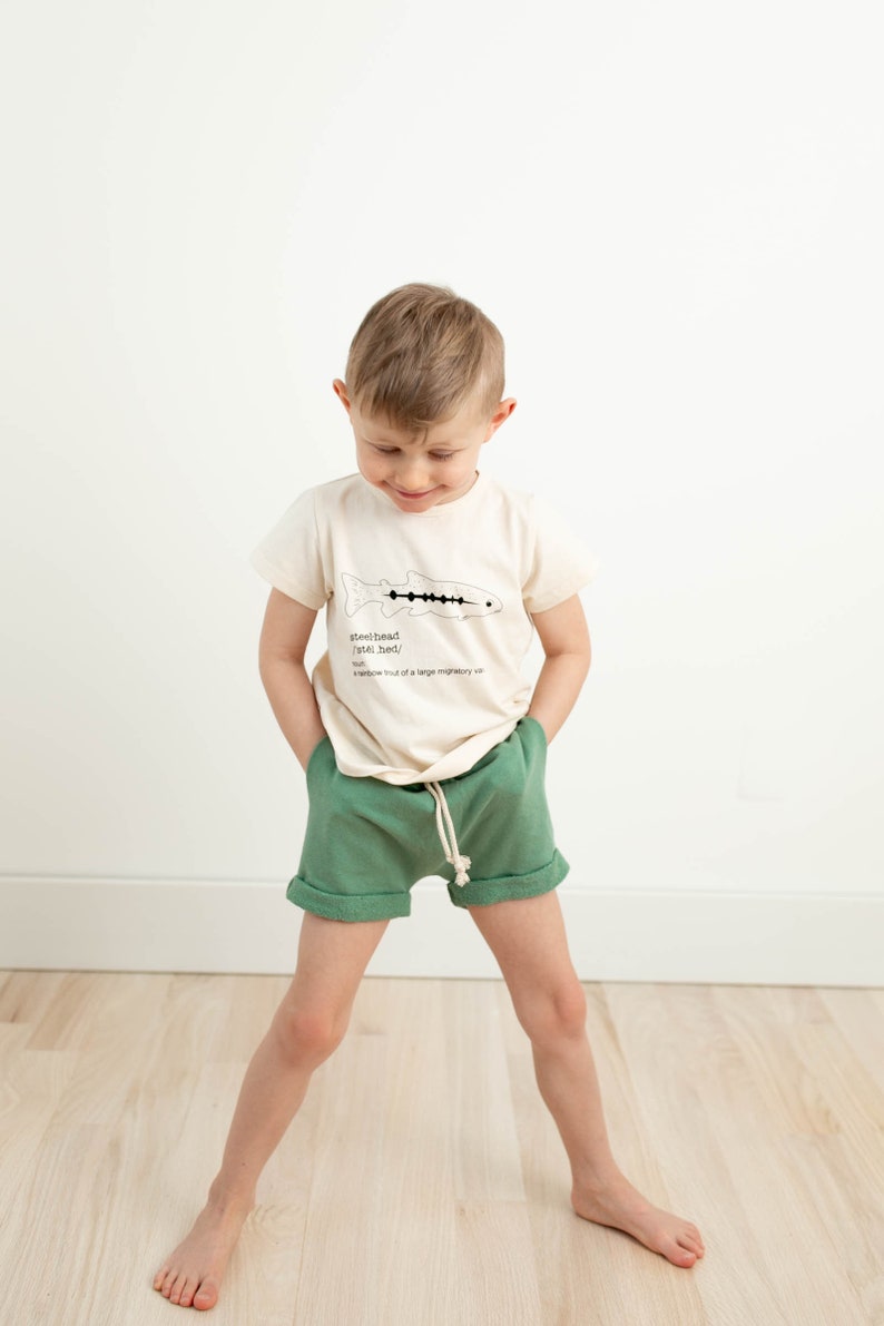 May include: A young boy wearing a cream colored t-shirt with a black and white illustration of a fish and the text "steelhead (steel-hed)" and green shorts.