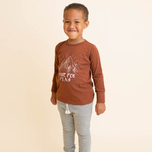 May include: A young boy wearing a brown long-sleeve shirt with a white mountain graphic and the text "MADE FOR PLAY". He is also wearing gray and white striped pants.