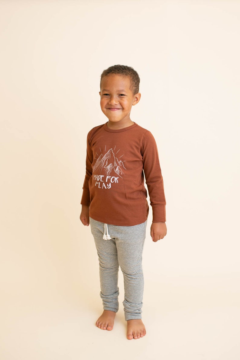 May include: A young boy wearing a brown long-sleeve shirt with a white mountain graphic and the text "MADE FOR PLAY". He is also wearing gray and white striped pants.