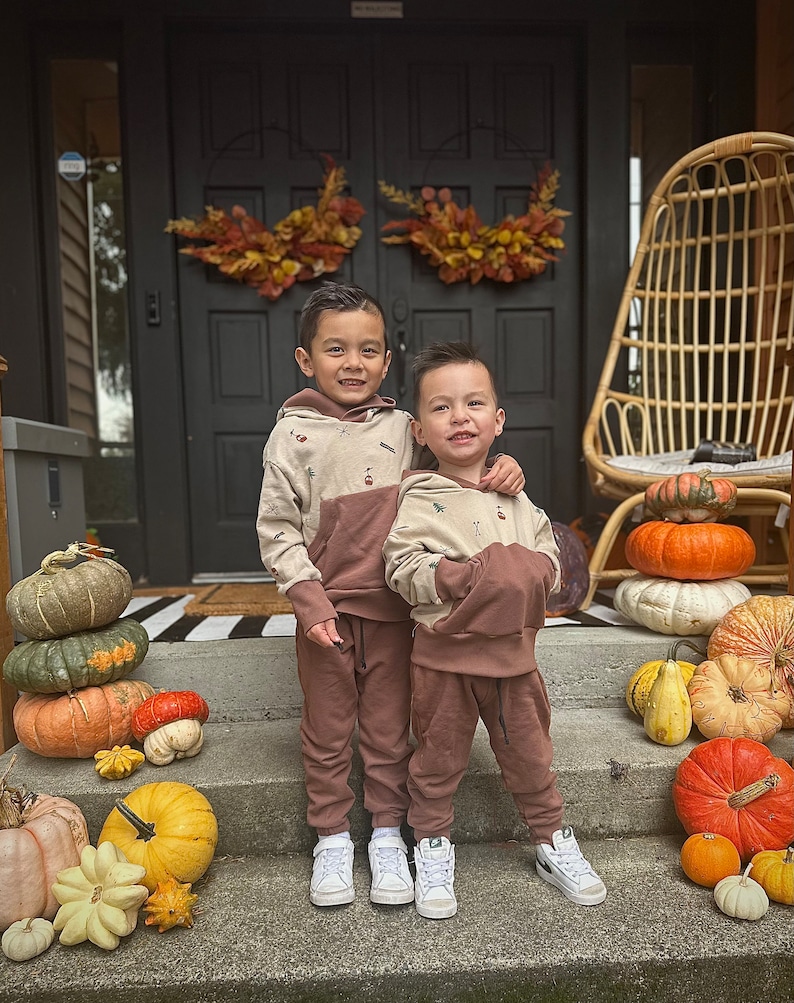 May include: Two young boys wearing brown and beige hoodies with a nature-themed print stand on a porch steps with a pile of pumpkins and gourds behind them.