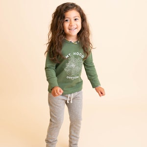 May include: A young person wearing a green long-sleeved shirt with a white graphic of Mt. Hood, Oregon. They are also wearing grey and white striped pants.