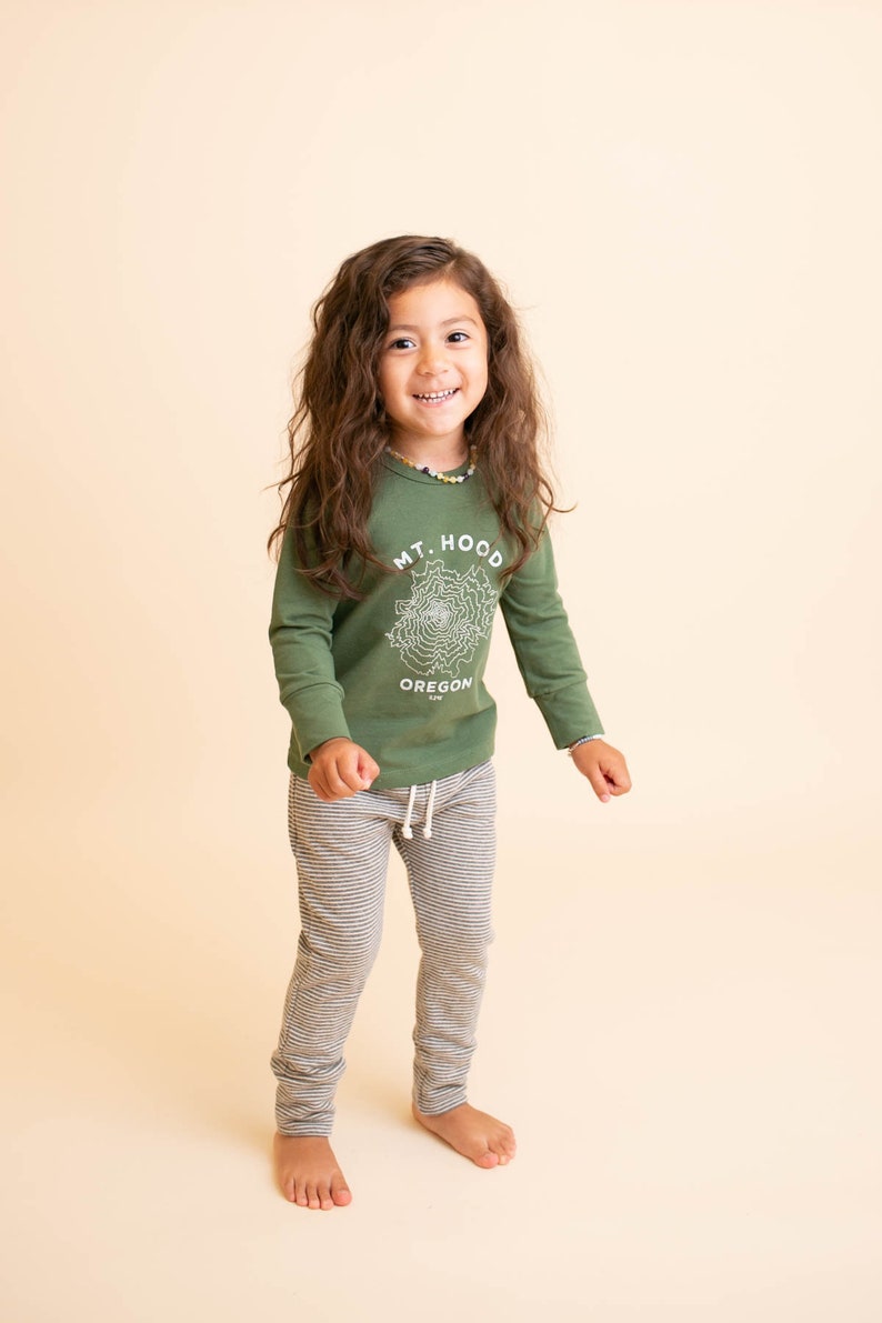 May include: A young person wearing a green long-sleeved shirt with a white graphic of Mt. Hood, Oregon. They are also wearing grey and white striped pants.