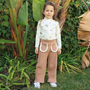 May include: A young person wearing a white long-sleeved shirt with a floral print and brown corduroy pants with white lace trim. The pants have two large pockets with white lace trim. The person is standing in front of a green leafy background.