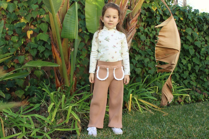 May include: A young person wearing a white long-sleeved shirt with a floral print and brown corduroy pants with white lace trim. The pants have two large pockets with white lace trim. The person is standing in front of a green leafy background.