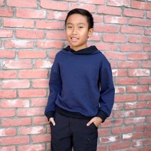 May include: A young person wearing a navy blue hooded sweatshirt with black trim and black cargo shorts. They are standing in front of a brick wall.