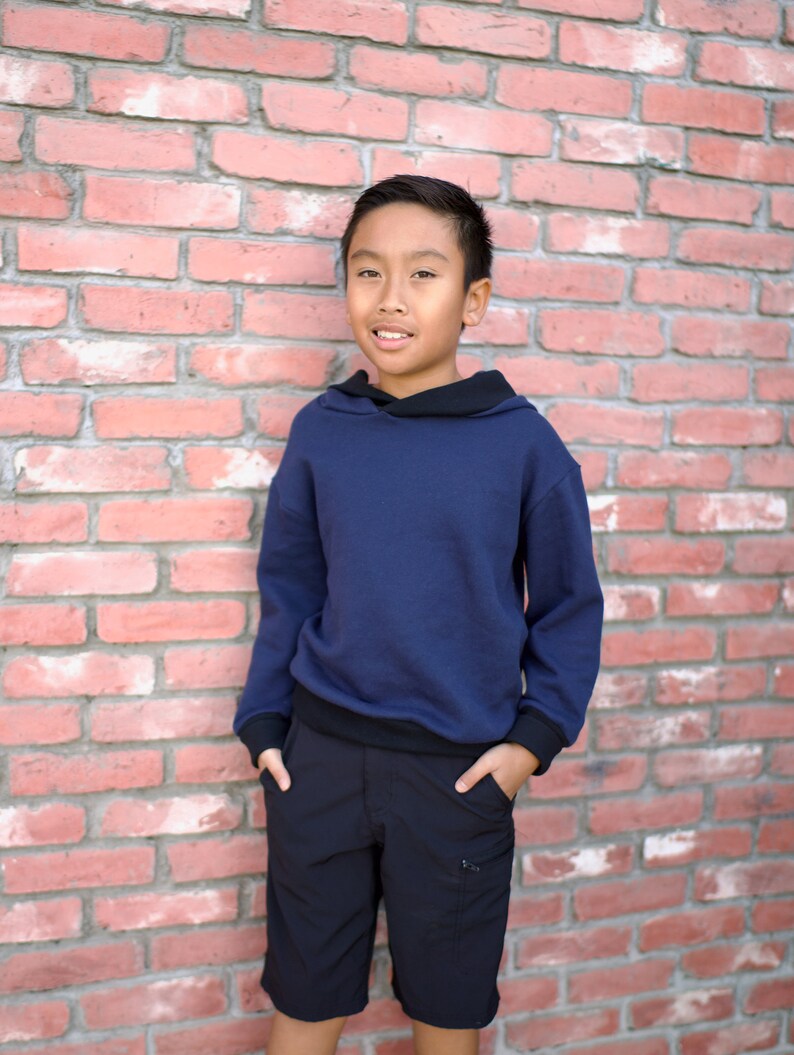 May include: A young person wearing a navy blue hooded sweatshirt with black trim and black cargo shorts. They are standing in front of a brick wall.