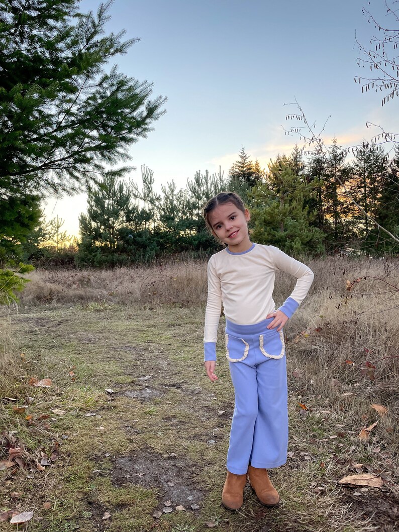 May include: A young person wearing a light beige long-sleeved shirt with blue pants that have white trim around the pockets. The pants are wide-legged and the person is standing on a path in a grassy area.