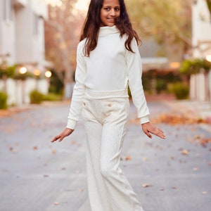 May include: A young person wearing a white turtleneck sweater and matching white pants with a cable knit pattern. They are standing on a paved street with fallen leaves.
