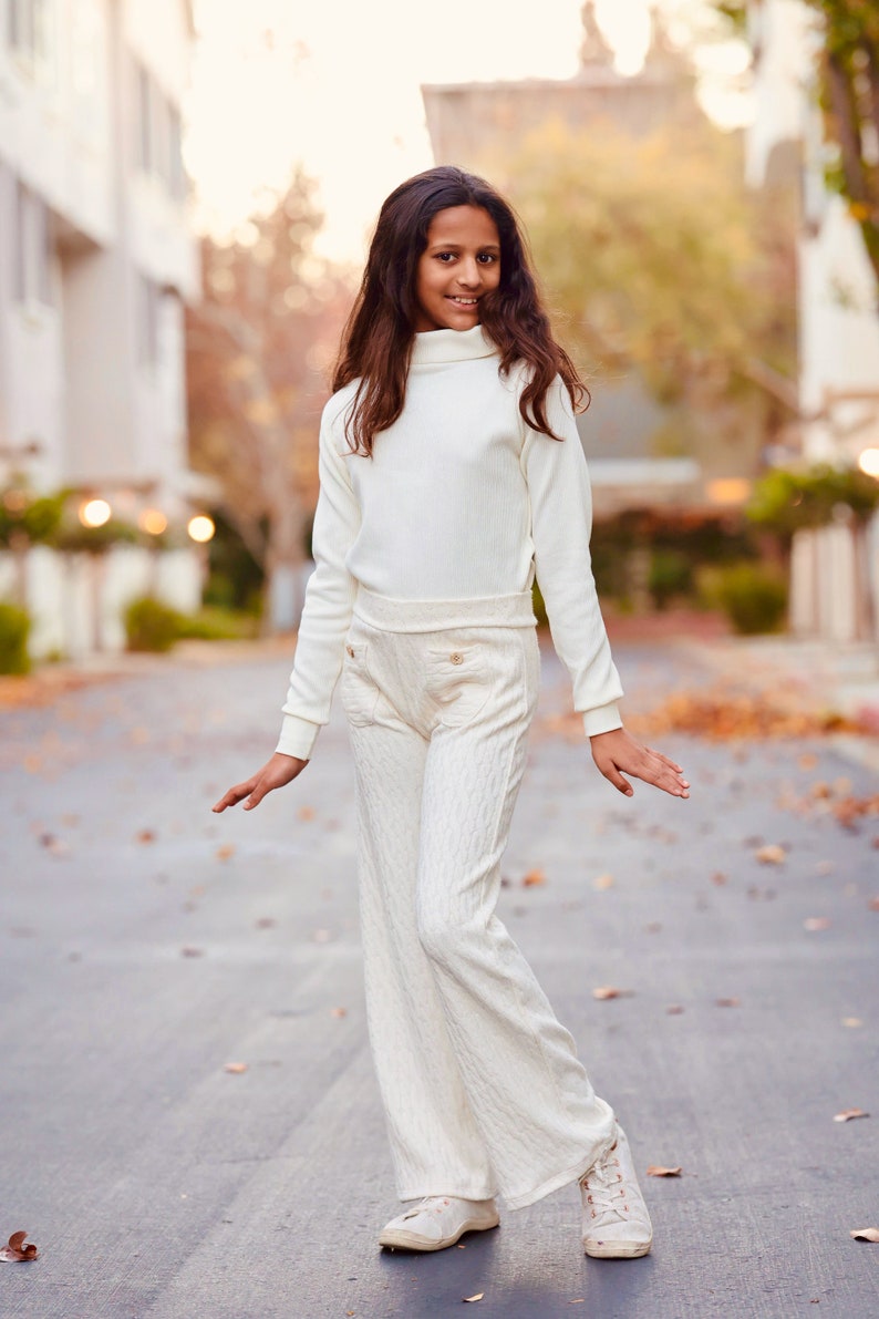 May include: A young person wearing a white turtleneck sweater and matching white pants with a cable knit pattern. They are standing on a paved street with fallen leaves.