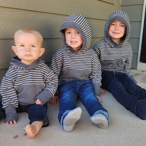 May include: Three young boys wearing striped hooded sweatshirts and jeans are sitting on a concrete porch. The boy on the left is wearing a gray and white striped hooded sweatshirt and blue jeans. The boy in the middle is wearing a gray and white striped hooded sweatshirt and blue jeans. The boy on the right is wearing a gray and white striped hooded sweatshirt and blue jeans.
