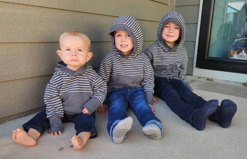 May include: Three young boys wearing striped hooded sweatshirts and jeans are sitting on a concrete porch. The boy on the left is wearing a gray and white striped hooded sweatshirt and blue jeans. The boy in the middle is wearing a gray and white striped hooded sweatshirt and blue jeans. The boy on the right is wearing a gray and white striped hooded sweatshirt and blue jeans.