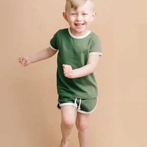 May include: A young boy wearing a green short-sleeved t-shirt with white trim and matching green shorts with white trim. He is smiling and running towards the camera.