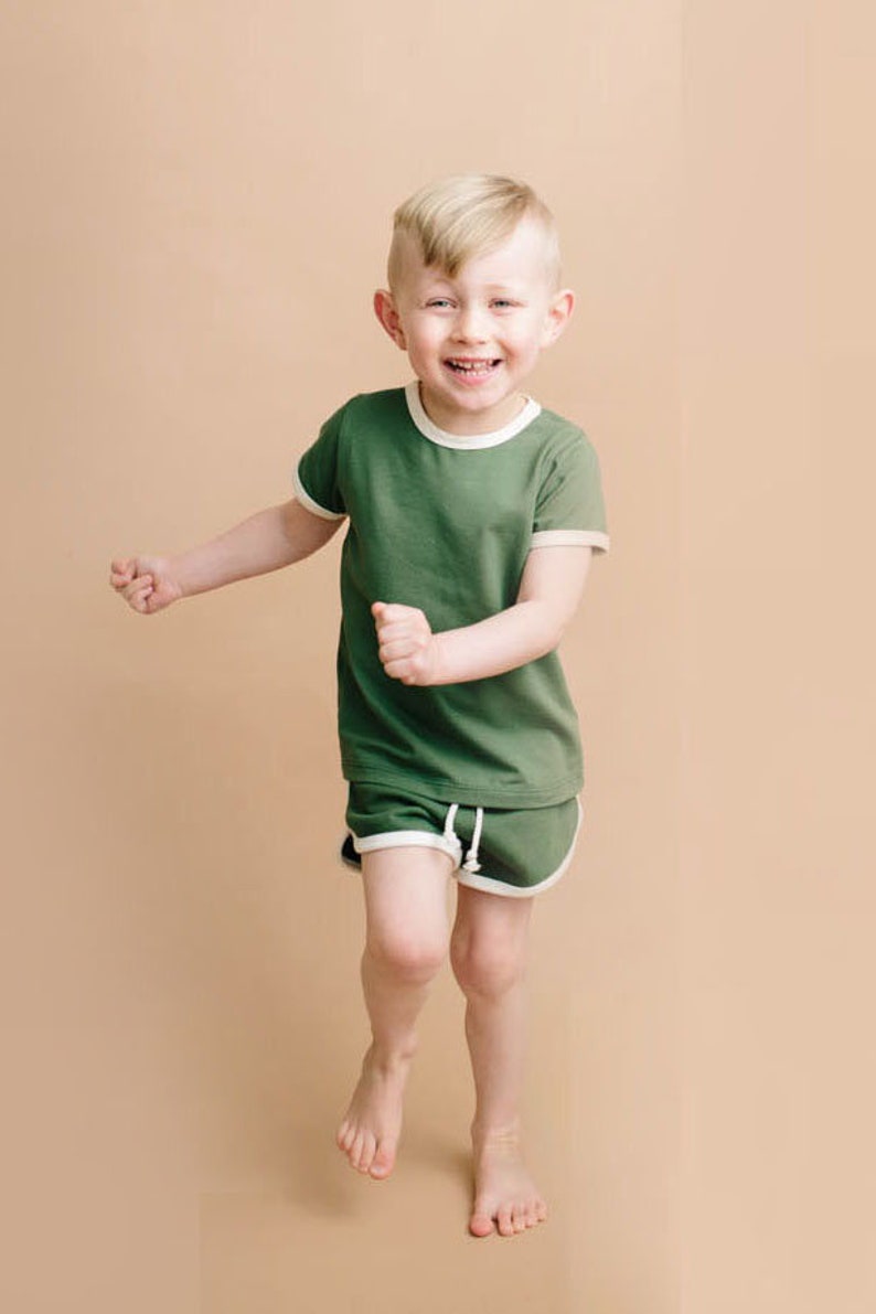 May include: A young boy wearing a green short-sleeved t-shirt with white trim and matching green shorts with white trim. He is smiling and running towards the camera.