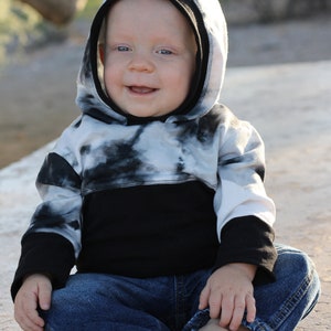 May include: A young child wearing a black and white tie-dye hoodie with a black band across the bottom. The child is wearing blue jeans and has their feet bare.