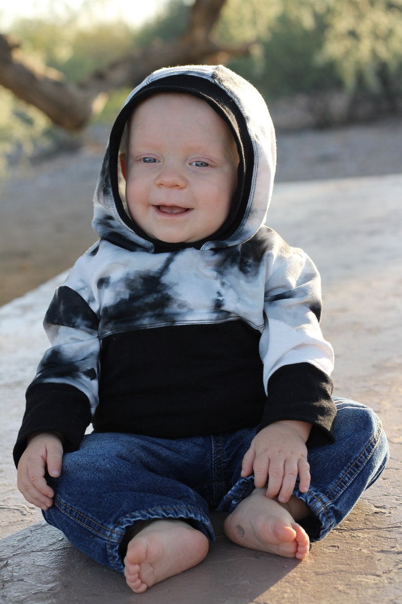 May include: A young child wearing a black and white tie-dye hoodie with a black band across the bottom. The child is wearing blue jeans and has their feet bare.