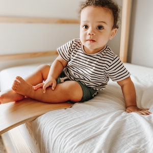 May include: A young child with brown curly hair sits on a white bed wearing a black and white striped shirt and blue denim shorts. The child is sitting on a wooden balance board.